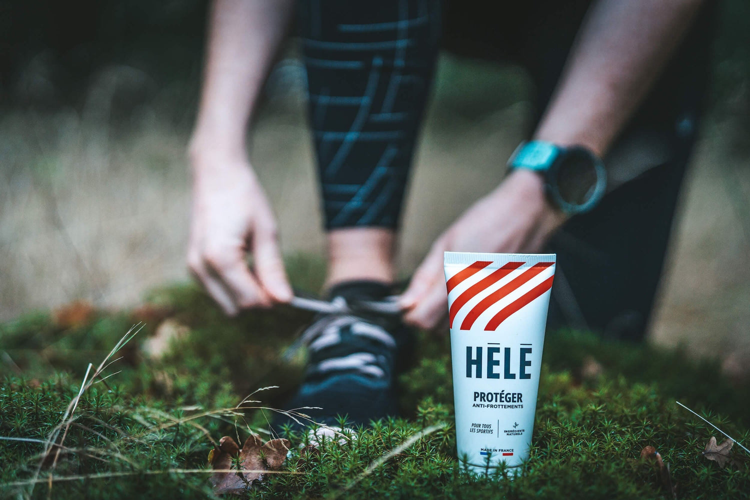 Gros plan sur un tube de crème anti frottements Protéger de la marque HELE posé dans la forêt avec un pied de coureuse refaisant ses lacets en arrière plan